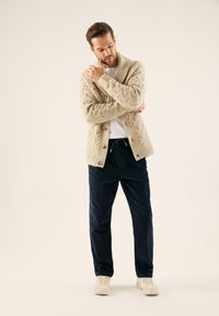 Beige kabelstrikket cardigan med store knapper, kombineret med marineblå fløjlsbukser og cremefarvede sneakers. Fokus på tekstur og farvekontrast.