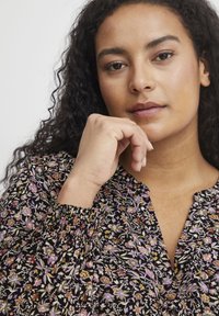 Melna ziedu blūze ar garnitūras kakla izgriezumu un garām piedurknēm. Iekļauj mazos purpura, dzeltenos un baltos ziedus uz tumša fona.