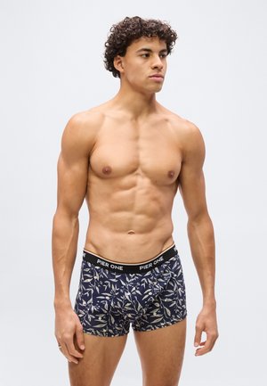 Uomo muscoloso con capelli ricci che indossa boxer blu navy con motivo a foglie bianche e fascia in vita nera con l'etichetta "Pier One", in piedi su sfondo bianco.
