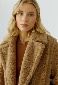 Blonde vrouw met groene ogen, draagt een bruin gebreid vest en een getextureerde camel-kleurige teddyjas, weergegeven vanaf de borst omhoog op een lichte achtergrond.
