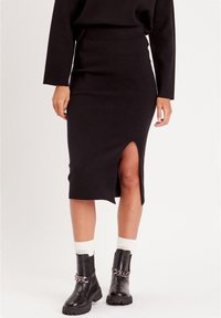 Jupe noire mi-longue avec une fente sur le côté, fabriquée en tissu doux. Associée à des bottines noires avec un accent en chaîne argentée.
