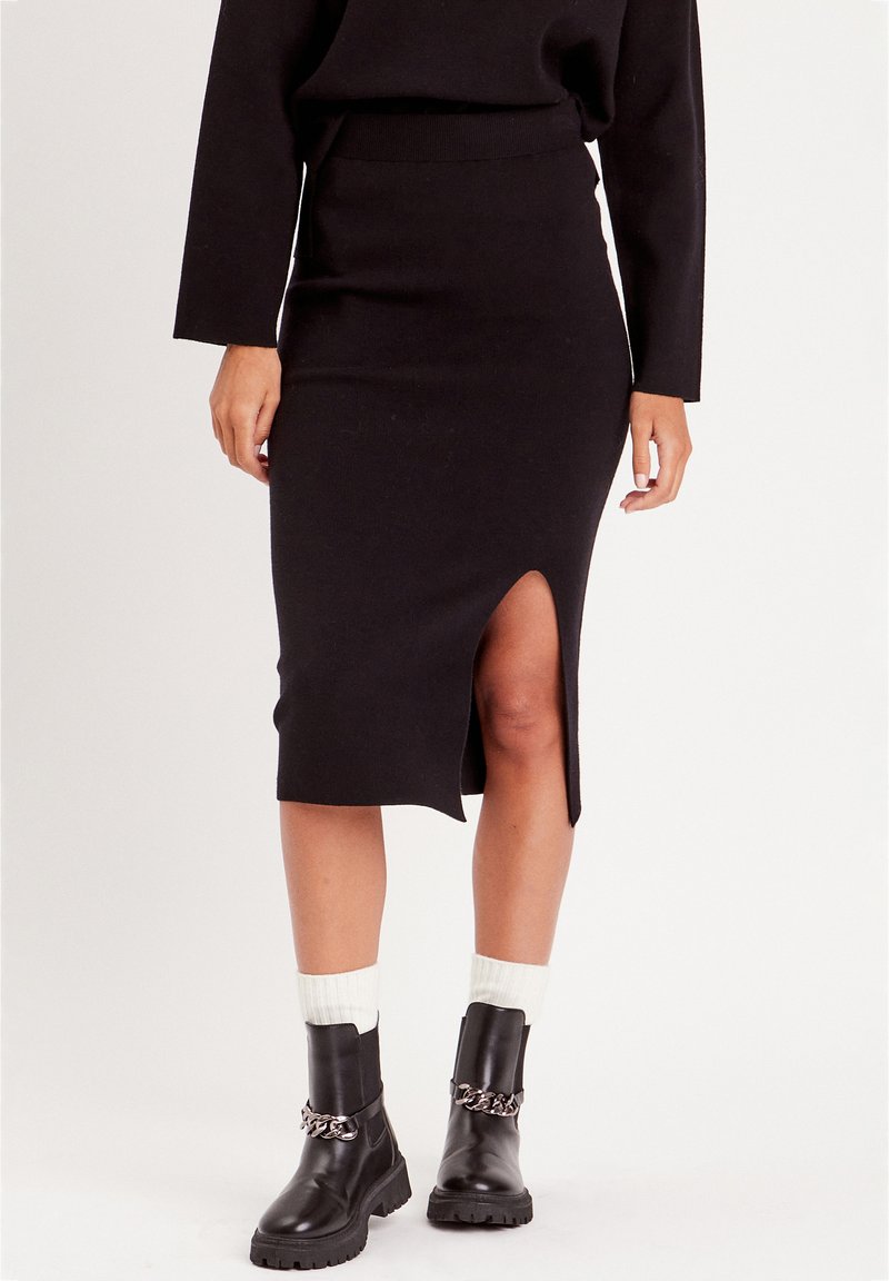 Jupe noire mi-longue avec une fente sur le côté, fabriquée en tissu doux. Associée à des bottines noires avec un accent en chaîne argentée.