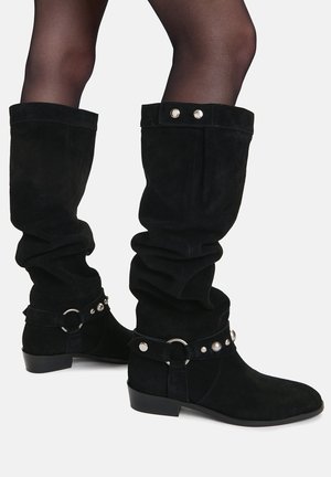 Frau trägt schwarze, velourslederne, lässige kniehohe Stiefel mit silbernen Nieten und Ringen, kombiniert mit durchsichtigen schwarzen Strumpfhosen vor weißem Hintergrund.