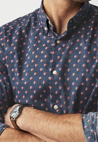 Man met een blauwe overhemd met een klein roze bloemenpatroon, met gekruiste armen en een zilveren horloge met een bruine leren band.