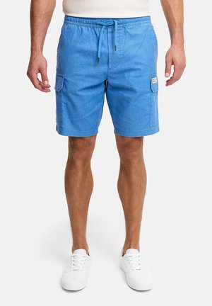 Mężczyzna ubrany w jasnoniebieskie szorty typu cargo z bocznymi kieszeniami, białe trampki i biały top, stoi na tle jednolitego tła.