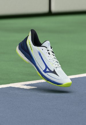 Scarpa sportiva bianca, blu e verde neon sospesa sopra un campo da tennis con superfici verde e blu e una linea di confine bianca.