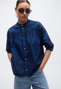 Dunkelblaues Jeanshemd mit hohem Kragen, gerafften Details, kurzen Ärmeln und Knopfleiste vorne, kombiniert mit hellblauen Jeans.