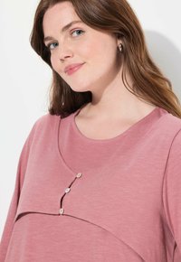 Maglia a maniche lunghe in morbido tessuto rosa, caratterizzata da un collo rotondo, design a strati e tre bottoni decorativi al centro davanti.