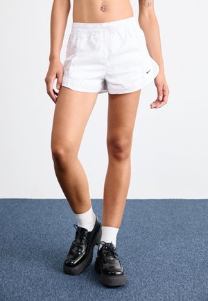Personne portant un short de sport blanc, des chaussettes blanches montantes et des chaussures à plateforme noires lacées, debout sur un tapis bleu.