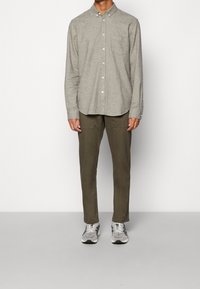 Graues Hemd mit Knöpfen aus strukturiertem Stoff und einer Brusttasche. Olivgrüne Hosen mit schmaler Passform. Graue und weiße Schnürschuhe.