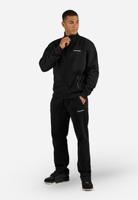 Zwarte zip-up jas en bijpassende broek van zachte stof, met voorzakken en witte logo-accenten. Model poseert tegen een effen achtergrond.