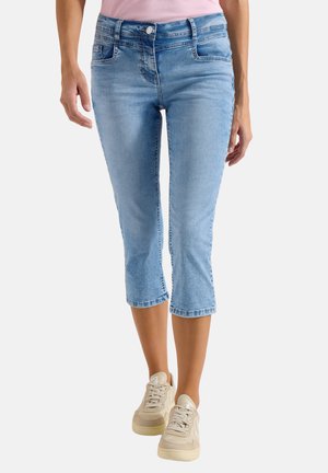 Frau trägt hellblaue, kürzere Jeans aus Denim und beigen Turnschuhe, steht vor einem einfarbigen Hintergrund.