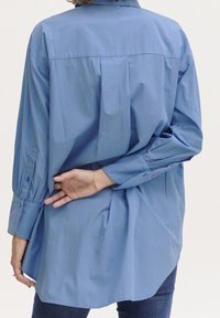 Camicia azzurra chiara con bottoni, maniche lunghe e orlo curvo. Presenta un colletto classico e dettagli in pieghe sul retro. Texture liscia.