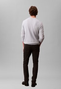 Light gray long-sleeve shirt with ribbed cuffs and a slightly rounded neckline, paired with dark jeans and black shoes. Rear view.