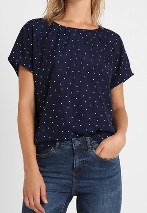 Kobieta ubrana w granatową bluzkę z krótkim rękawem w białe kropki, wsuniętą w niebieskie dżinsy, z jedną ręką w kieszeni.