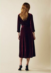 Burgundin samettimekko, jossa on puolipitkät hihat, vyötäröä korostava malli ja laskeutuva hame, vyö ja käärityt hihat, yhdistetty mustiin korkokenkiin.