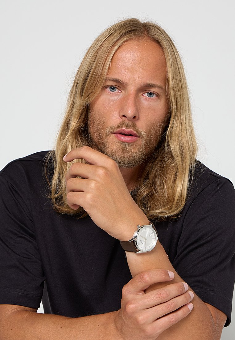 Silberne Analog-Uhr mit weißem Zifferblatt und braunem Lederband, am Handgelenk getragen. Das Modell hat lange Haare und trägt ein schwarzes Shirt.