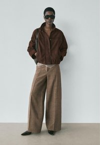 Braune Wildlederjacke mit hohem Kragen, beiges Spitzenoberteil und weite braune karierte Hose. Schwarze spitze Schuhe. Model trägt Sonnenbrille.