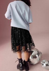 Lichtblauwe oversized t-shirt, zwarte kanten rok met pailletten en chunky zwarte laarzen; staande naast spiegelende discobollen op een roze oppervlak.