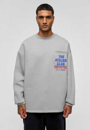 Lichtgrijs sweatshirt met een ronde hals, grote zak aan de linkerkant en vetgedrukte tekst in blauw en rood. Voorzien van geribde boorden en zoom.