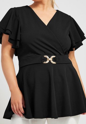 Kvinde iført en sort omslagstop med lagdelte flutterærmer og et bælte med guldlås, kombineret med hvide bukser.