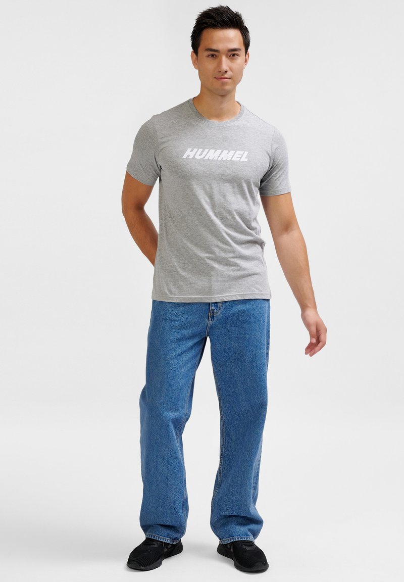 Grå t-shirt med "HUMMEL" logo, parret med løstsiddende blå denim jeans og sorte sneakers, stående foran en ensfarvet baggrund.