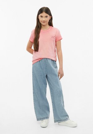 Junges Mädchen mit langen braunen Haaren trägt ein pinkes T-Shirt, weite hellblaue Jeans und weiße Turnschuhe, steht vor einem schlichten weißen Hintergrund.