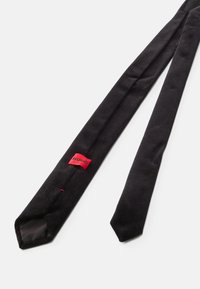 Corbata negra texturizada con un extremo puntiagudo y una etiqueta roja "HUGO". Tela suave, diseño sólido, sin patrones ni adornos.