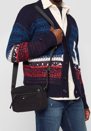 Sac à bandoulière en suède noir avec un haut zippé, une poche plate à l'avant et une bandoulière amovible. Surface texturée avec des accents élégants.