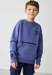 Lilla kapuutsärkk, millel on ees lukuga tasku, soonikkoes varrukad ja logo rinnal. Kandev on tumesinised cargo-püksid. Sile tekstuur.