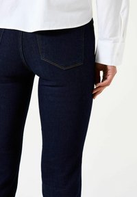 Jean en denim bleu foncé à coupe près du corps, avec des coutures contrastantes et deux poches arrière. Chemise blanche boutonnée partiellement visible.