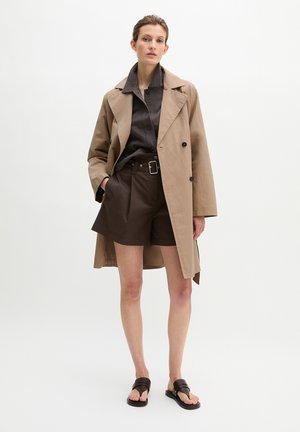 Woman wearing a beige trench coat, dark brown button-up shirt, matching belted shorts, and dark brown sandals, standing with one hand in pocket.