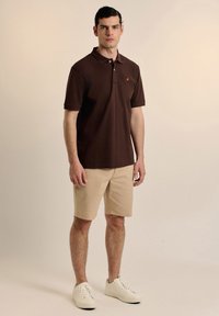 Camisa polo marrón oscuro con cuello y mangas cortas, combinada con pantalones cortos beige y zapatillas blancas. Presenta un pequeño logo bordado.