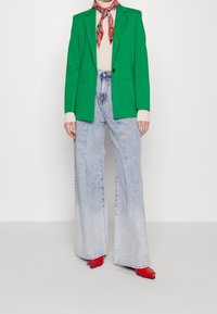 Blazer strutturato verde, top crema, jeans a vita alta blu chiaro a zampa d'elefante e scarpe rosse con tacco. La sciarpa presenta un motivo colorato.