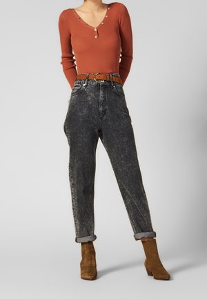 Kvinde iført rustfarvet, ribstrikket langærmet top med knappedetaljer, sort højtsiddende jeans med syrevaskeffekt, brun bælte og brune ruskindsstøvler til anklerne.