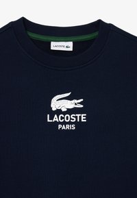 Sweatshirt bleu marine avec col côtelé, arborant le logo Lacoste blanc et un graphique de crocodile. Le tissu a une texture lisse et douce.