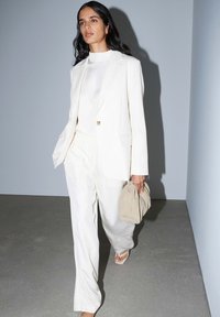 Donna che cammina al chiuso indossando un completo completamente bianco e sandali, con una borsa clutch morbida beige contro pareti grigie semplici.