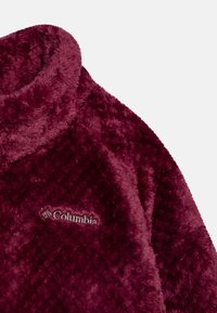 Pullover in velluto borgogna con colletto alto, texture morbida e motivo trapuntato. Presenta un piccolo logo Columbia ricamato sul petto.