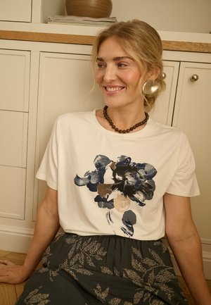 Femme aux cheveux blonds portant un t-shirt blanc à motif floral et une jupe sombre avec des motifs de feuilles, assise sur un sol en bois devant des armoires couleur crème.