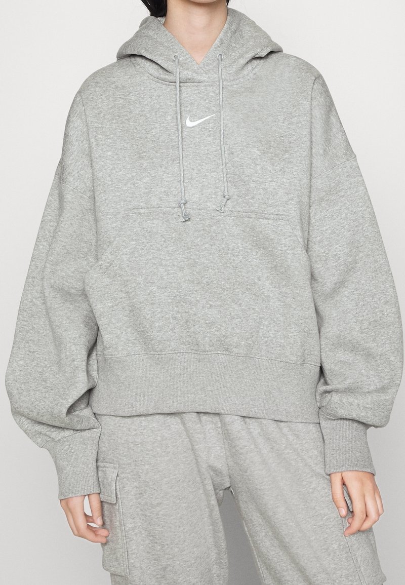 Personne portant un sweat à capuche Nike gris clair avec cordons et un pantalon assorti avec une poche latérale sur un fond uni.