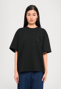 Μαύρη κοντομάνικη μπλούζα T-shirt με στρογγυλή λαιμόκοψη και κεντητό σχέδιο στην αριστερή πλευρά. Λαμπερός ύφασμα με χαλαρή εφαρμογή.