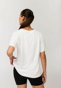 Frau mit dunklen Haaren im Pferdeschwanz, die ein lockeres weißes T-Shirt und schwarze Biker-Shorts trägt, steht mit einer Hand auf der Hüfte und schaut weg.