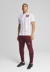 Camisola atlética branca com riscas verticais em bordeaux e logótipo, combinada com calças bordeaux com detalhes em dourado e marca. Sapatos de futebol pretos.