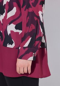 Blusa a maniche lunghe nelle tonalità di bordeaux e crema con motivo floreale astratto. Presenta un polsino attillato con dettaglio a bottone. Tessuto morbido.