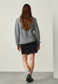 Woman with long brown hair walking away, wearing a gray sweater, black skirt, gray socks, and black shoes on a gray floor.