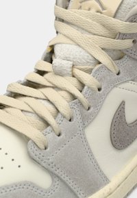 Κοντινό πλάνο από σουέτ αθλητικά παπούτσια σε μπεζ και γκρι χρώμα με κρεμ κορδόνια και τρυπητό μπροστινό μέρος, που αναδεικνύει τις υφές των υλικών και το λογότυπο της Nike.