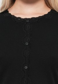 Svart ribbet cardigan med bølget blondestoff og seks knapper foran som lukking. Glatt tekstur med subtil detaljering.