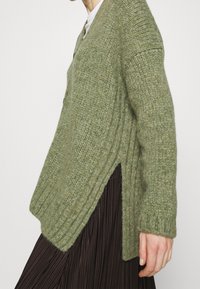 Green knitted cardigan with a ribbed texture, v-neck, and side slits. Long sleeves and lightweight material, paired with a pleated brown skirt.