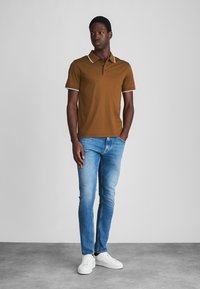 Braunes Poloshirt mit weißen Akzenten, kombiniert mit hellblauen Jeans und weißen Sneakers. Das Shirt verfügt über eine Zwei-Knopf-Leiste und eine glatte Textur.