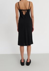 Vestido negro de midi con tirantes finos, abertura en la espalda en forma de V y detalle de lazo. Textura suave, tela blanda, cae justo por encima de los tobillos.